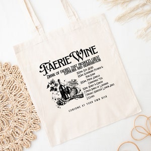 May include: Cream-colored tote bag with a black graphic and text that reads "Faerie Wine." The graphic includes a wine bottle, fruit, and text listing ingredients. The bag has a simple design with two handles.