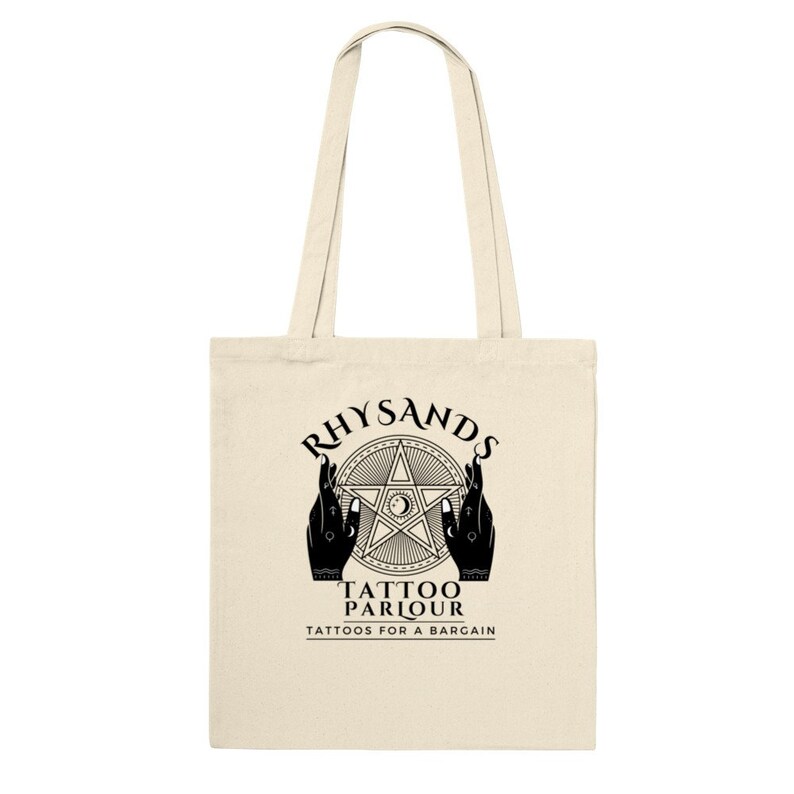 May include: A beige tote bag with black text and a graphic design. The text reads "Rhysands Tattoo Parlour Tattoos for a Bargain". The graphic is a five-pointed star with a crescent moon and eye inside, surrounded by two hands.