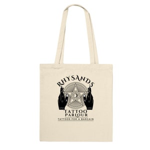 May include: A beige tote bag with black text and a graphic design. The text reads "Rhysands Tattoo Parlour Tattoos for a Bargain". The graphic is a five-pointed star with a crescent moon and eye inside, surrounded by two hands.