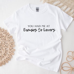May include: White t-shirt featuring the phrase "YOU HAD ME AT Enemies to Lovers" in black. The shirt is made of a soft material and is displayed on a white surface. Woven decorations are visible in the background.