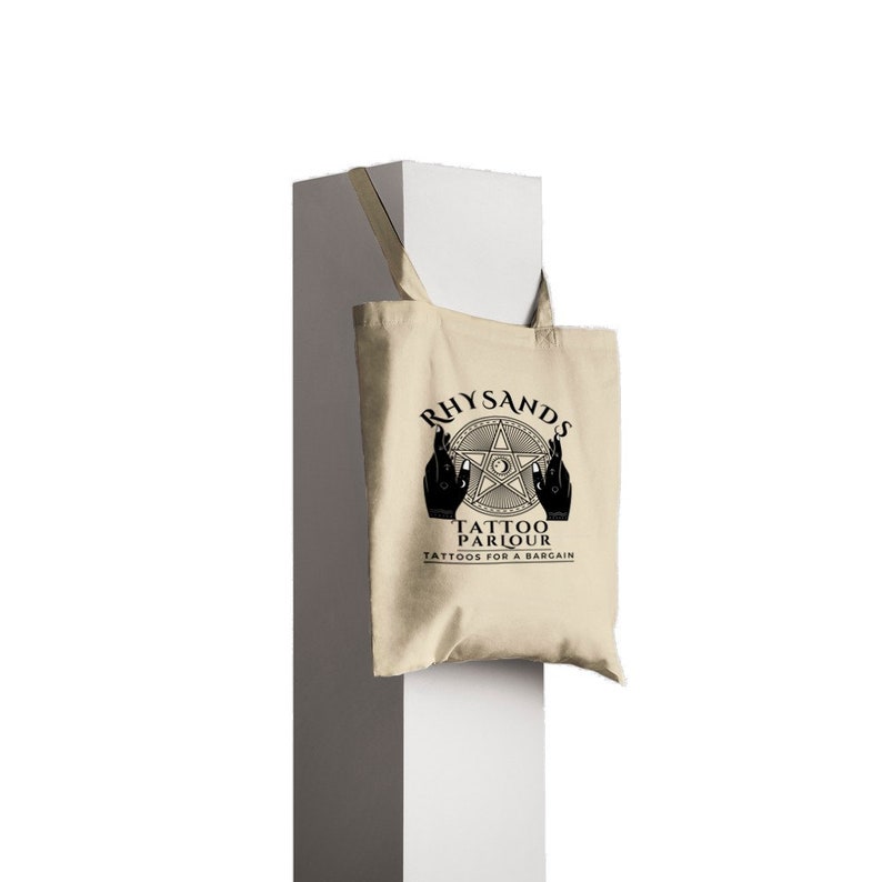 May include: A beige tote bag with black handles and a black graphic design. The graphic features a five-pointed star with a circle in the center, two hands reaching towards the star, and the text "Rhysands Tattoo Parlour Tattoos for a bargain".