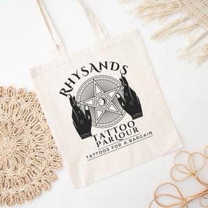 May include: A white canvas tote bag with black text and a graphic design. The text reads "Rhysands Tattoo Parlour Tattoos for a Bargain". The graphic is a five-pointed star with two hands holding it up.