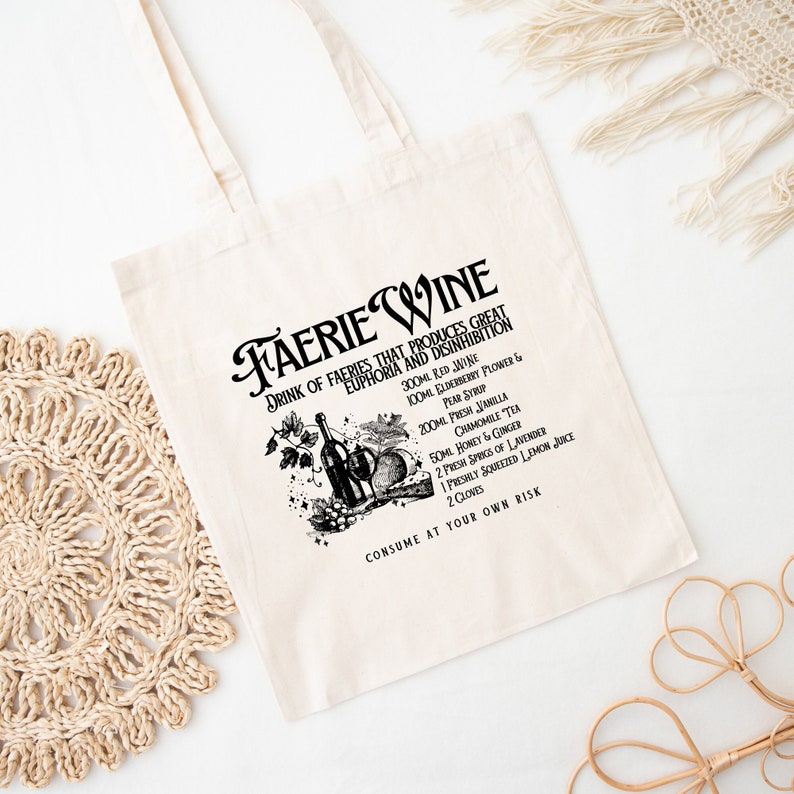 May include: Cream-colored tote bag with a black graphic and text that reads "Faerie Wine." The graphic includes a wine bottle, fruit, and text listing ingredients. The bag has a simple design with two handles.