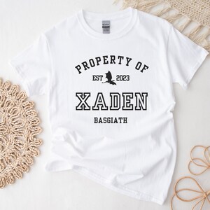 May include: White t-shirt with black text that reads "PROPERTY OF EST 2023 XADEN BASSGIATH" with a black silhouette of a dragon above the name.