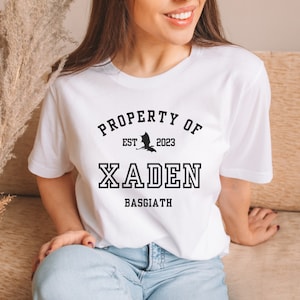 May include: White t-shirt with black text that reads "PROPERTY OF EST 2023 XADEN BASSGIATH" with a black silhouette of a dragon above the text.