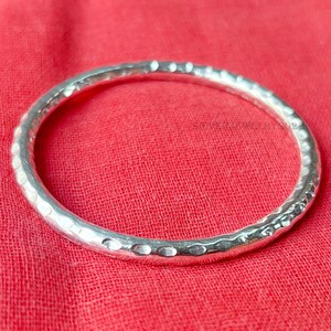 Könnte beinhalten: Ein gehämmertes Silberarmband. Das Armband hat eine strukturierte Oberfläche mit kleinen, unregelmäßigen Vertiefungen. Das Armband ist kreisförmig und liegt auf einem roten Stoff. Das Armband ist ein Schmuckstück.