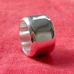 Könnte beinhalten: Ein polierter Silberring mit breitem Band. Der Ring hat eine glatte, reflektierende Oberfläche und eine abgerundete Form. Der Ring ist vor rotem Hintergrund platziert.