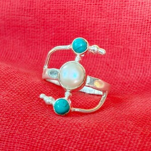 Könnte beinhalten: Ein silberner Ring mit einer weißen Perle und zwei türkisfarbenen Steinen. Der Ring ist mit einem gebogenen Band und einem filigranen, durchbrochenen Design gestaltet.