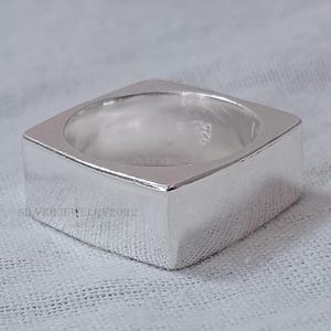 Könnte beinhalten: Ein quadratischer Silberring mit glatter, polierter Oberfläche. Der Ring hat ein quadratisches Band und eine quadratische Oberseite. Der Ring ist auf der Innenseite des Bandes mit der Nummer "925" gekennzeichnet.