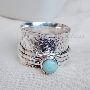 Könnte beinhalten: Ein silberner Spinner-Ring mit floralem Muster und einem runden, hellblauen Edelstein. Der Ring hat ein strukturiertes Band und einen sich drehenden Außenring. Der Edelstein ist in einer Lünettenfassung gefasst.