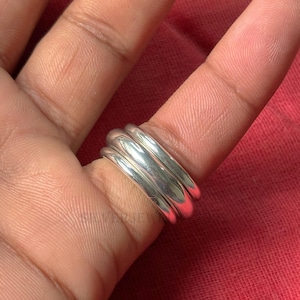 Könnte beinhalten: Ein silberner Ring mit drei gestapelten Bändern. Der Ring ist glatt und glänzend und reflektiert das Licht. Der Ring wird auf einem Finger vor rotem Hintergrund gezeigt. Der Schmuck ist wahrscheinlich ein Modeaccessoire.