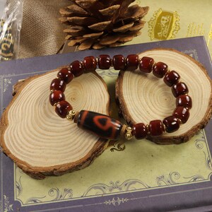May include: A beaded bracelet featuring dark red, rectangular beads and a central cylindrical bead with a black and red pattern. The bracelet is accented with gold-colored spacers and is displayed on a wooden surface.
