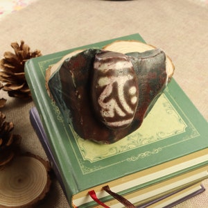 May include: A dzi bead, featuring a white and brown pattern, is set in a dark brown stone. The stone rests on a stack of books with green covers. The bead's design is a prominent focal point, showcasing intricate details.