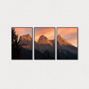 May include: A triptych of framed mountain landscape art. The artwork features three separate black framed prints of mountain peaks under a vibrant orange and pink sunset sky. The mountains are brown and gray, with some green trees in the foreground.
