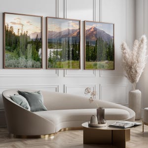 May include: Three framed landscape prints depicting mountain scenery with trees and a lake. The frames are a warm brown, and the artwork is displayed above a curved beige sofa with gray pillows. A coffee table and decorative vase are in front.