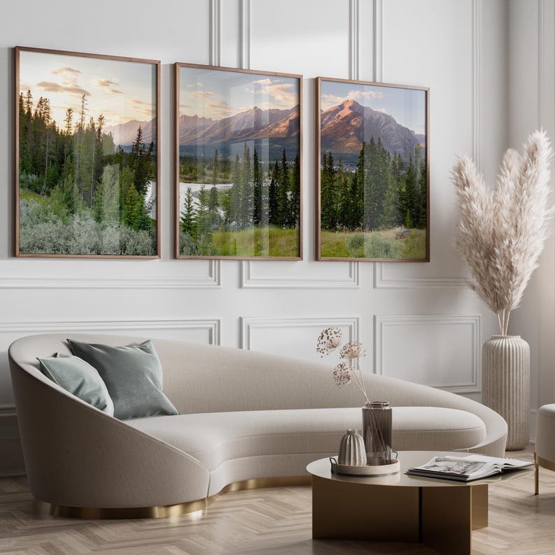 May include: Three framed landscape prints depicting mountain scenery with trees and a lake. The frames are a warm brown, and the artwork is displayed above a curved beige sofa with gray pillows. A coffee table and decorative vase are in front.