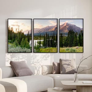 May include: Three framed landscape prints showing mountain and forest scenes. The artwork features a river, trees, and mountains under a sunset sky. The frames are black, and the colour palette includes greens, blues, and golds.