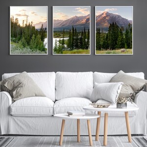May include: Three framed landscape prints featuring mountain and forest scenes. The artwork showcases a lake, trees, and mountains under a sunset sky. The prints are displayed above a white sofa with neutral-toned pillows and a coffee table.
