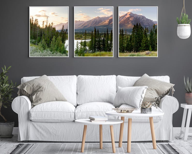 May include: Three framed landscape prints featuring mountain and forest scenes. The artwork showcases a lake, trees, and mountains under a sunset sky. The prints are displayed above a white sofa with neutral-toned pillows and a coffee table.