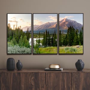 May include: A triptych canvas print featuring a scenic mountain landscape with a river and lush green trees. The artwork is framed in black and displayed on a dark wooden cabinet. The sky is a mix of blue and orange hues.