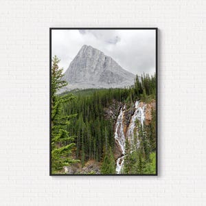 May include: A framed print of a mountain landscape with a waterfall. The mountain is grey, with a waterfall flowing down its side. Dense green trees surround the base of the mountain and waterfall, under a cloudy sky.