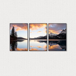 May include: Three framed landscape prints. Each features a serene lake scene with mountain reflections. The sky is filled with vibrant orange and pink clouds, reflecting in the calm water. A tall evergreen tree is visible in one frame.