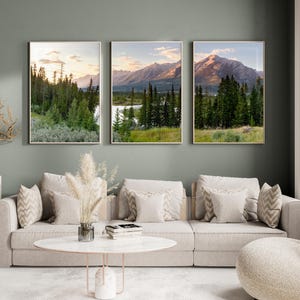 May include: Three framed landscape prints depicting mountain scenery with trees and a river. The prints are displayed above a beige sofa with decorative pillows. The room has a neutral color palette with a modern aesthetic.