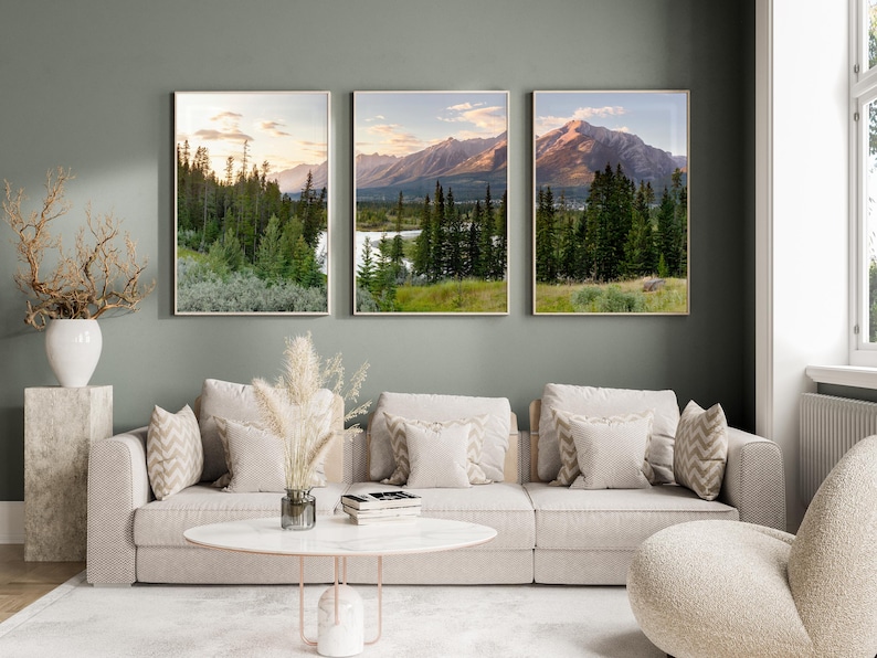 May include: Three framed landscape prints depicting mountain scenery with trees and a river. The prints are displayed above a beige sofa with decorative pillows. The room has a neutral color palette with a modern aesthetic.