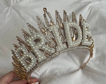 Op de afbeelding: Gouden kroon met het woord "BRIDE" gespeld in witte parels. De kroon is versierd met strass.