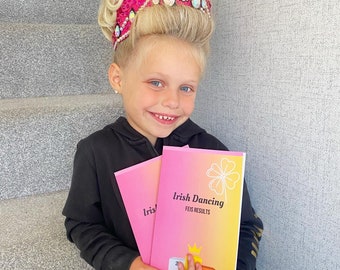 Op de afbeelding: Een jong meisje in een zwart jack en een roze kroon met strass houdt twee roze en gele brochures vast. De brochures hebben de tekst "Irish Dancing FEIS RESULTS" en een klavertjevier.