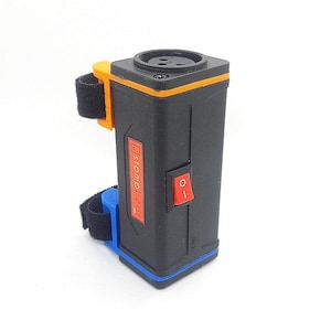 Puede incluir: Caja de efectos negra con ribete naranja y un interruptor rojo de encendido/apagado. La caja de efectos tiene una correa negra y un conector azul.