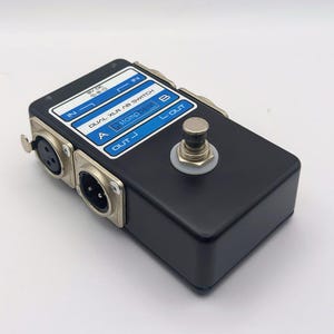May include: A black stompbox with silver XLR connectors and a silver footswitch. The box has a white and blue label that reads "DUAL XLR AB SWITCH" and "stompbox". The box has "IN", "OUT", and "A" and "B" labels.