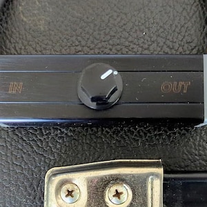 May include: A black audio device with a central knob and the words "IN" and "OUT" printed on the surface. The device has gold-colored accents and is connected to black cables. The device is set against a textured black surface.