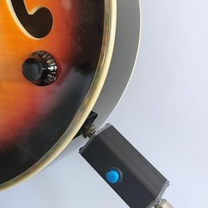 Puede incluir: Primer plano de una guitarra acústica con acabado sunburst, con perillas negras y un dispositivo rectangular negro con un botón azul conectado a la guitarra a través de un cable.