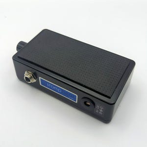 May include: A black stompbox pedal with a textured surface and a small blue label that reads "stompbeat.com". The pedal has a jack input, a power input, and a knob. The pedal is rectangular and has rounded edges.