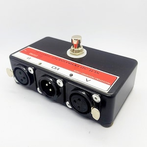 Może przedstawiać: Czarna skrzynka przełącznika audio XLR z trzema żeńskimi złączami XLR, czerwoną etykietą z napisem "PHANTOM POWERED 15V/105" i czarnym przełącznikiem nożnym ze srebrnym pokrętłem. Etykieta zawiera również napis "XLR 48V PERMANENT SWITCH".