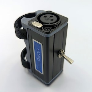 Può includere: Un dispositivo audio rettangolare nero e grigio dotato di una porta di ingresso XLR montata superiormente con un pulsante di rilascio argentato. Il lato presenta un'etichetta blu con il testo stompbeat.com e un interruttore a levetta in metallo.