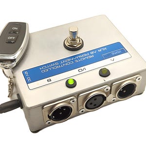Può includere: Un interruttore permanente a controllo remoto argentato con due connettori XLR, una luce LED verde e un telecomando nero con pulsanti ON e OFF. L'interruttore è etichettato "REMOTE CONTROLLED XLR A8 PERMANENT SWITCH 12V DC".
