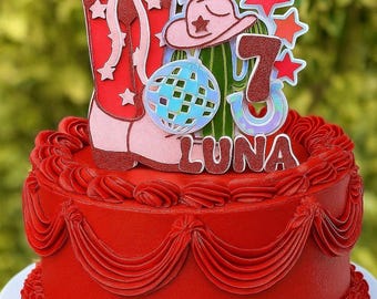 May include: A red frosted cake with a decorative top featuring a cowboy boot, stars, a cowboy hat, a disco ball, and the number 7. The cake has the name "LUNA" on it. The cake is on a white cake board.