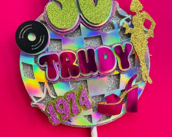 May include: A glittery 50th birthday cake topper with the name "Trudy" and the year "1974". The design includes a disco ball, a record, a silhouette of a woman, and a platform shoe. The topper is on a purple and white striped stick.