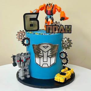 May include: A blue birthday cake decorated with Transformers-themed toys and decorations. The cake features an orange robot figure, a yellow car, and a silver Transformers logo. The number 6 and a name are also displayed.