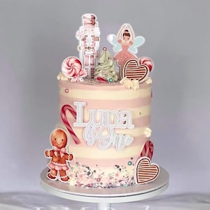 May include: A celebratory cake with pink and white stripes, decorated with a gingerbread person, a nutcracker, a ballerina, and candy. The cake is adorned with the words "Luna is One". Sprinkles and heart-shaped biscuits add to the festive theme.