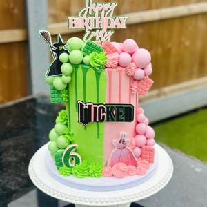May include: A two-tiered cake with green and pink frosting, decorated with a witch, the word "Wicked", and the text "Happy Birthday Emily". The cake is adorned with green and pink candies and sits on a white cake stand.