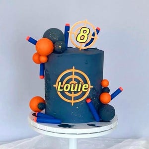 May include: A navy blue birthday cake decorated with orange and blue toy darts and spheres. The cake has a target design with the name "Louie" and the number "8" on top, perfect for a child's birthday.