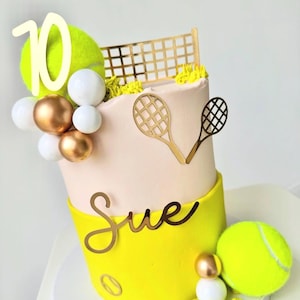 May include: A tennis-themed cake with a yellow and pink colour scheme. The cake is decorated with tennis balls, rackets, and a net. The name "Sue" and the number "10" are also featured in gold. The cake is a two-tiered design.