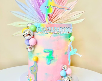 May include: A colorful birthday cake with a gymnastics theme. The cake is decorated with a rainbow-colored frosting, a number 7, and gymnast figures. The cake is adorned with disco ball accents and a topper that reads "ELORALELUISE".