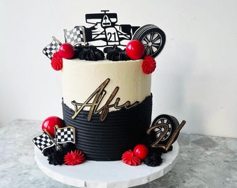 May include: A two-tiered cake with black and white frosting, decorated with a race car theme. The cake features a race car topper, checkered flags, red spheres, and the name "Afrie" in gold script. The number "21" is also displayed.