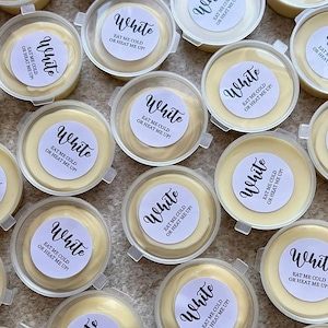 May include: An aerial view of numerous small, clear plastic pots filled with a creamy, off-white substance. Each pot has a white label with the word "White" and the text "EAT ME COLD OR HEAT ME UP!" in black. The pots are arranged closely together.