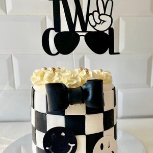 May include: A two-tiered cake with a black and white chequered pattern. The cake is decorated with a black bow, smiley face cutouts, and a topper that reads "TWO COOL" with sunglasses and a peace sign. The top is frosted with white buttercream and gold sprinkles.