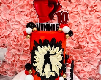 May include: A red and orange birthday cake with boxing theme. The cake features a silhouette of a boxer, a red boxing glove topper, and the name "Vinnie" with the number "10". The cake is decorated with black, white, and gold accents.